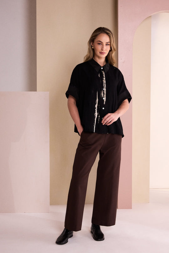 A woman modeling a black short-sleeve button-up shirt with a white vertical graphic print down the front, paired with dark brown wide-leg trousers and black shoes, standing in a minimalist studio with beige and pink architectural panels.