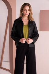A woman stands indoors wearing a black satin-like button-front jacket over a muted olive top and black wide-leg pants. The setting has soft pink and beige walls with architectural lines, and no visible brand marks are present.