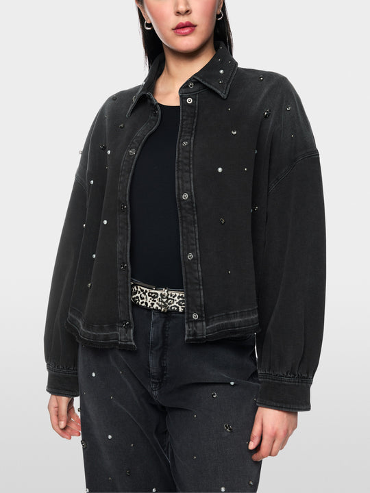 A woman wearing a black washed denim jacket adorned with small silver rhinestone studs, styled over a black top and dark jeans with matching rhinestone details, against a plain white background.