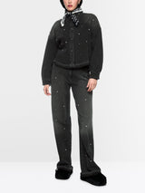 A model wearing a black denim jacket and matching wide-leg jeans, both embellished with small silver rhinestone studs, styled with a black bandana, black hat, and black fuzzy slippers against a light gray studio background.