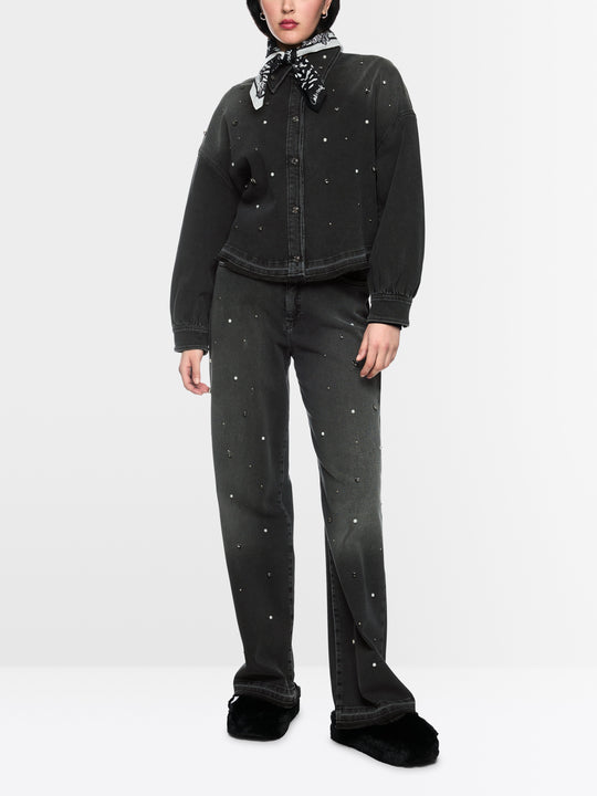 A model wearing a black denim jacket and matching wide-leg jeans, both embellished with small silver rhinestone studs, styled with a black bandana, black hat, and black fuzzy slippers against a light gray studio background.