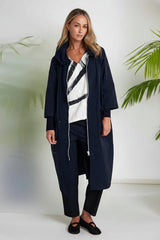 A woman modeling a long navy blue hooded coat over a white top with black diagonal stripes, paired with dark trousers and black flats, set against a white studio background with soft green plant shadows.