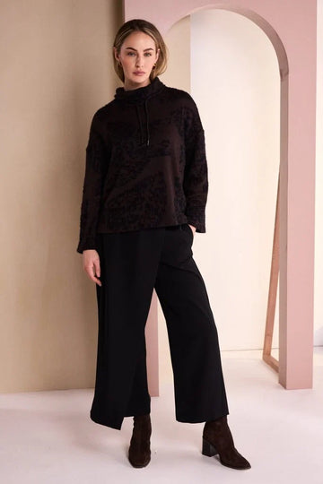 A woman modeling a dark brown textured knit mock-neck top and black wide-leg trousers with brown ankle boots, standing in a minimalist studio with beige and pink arches.