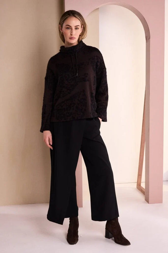 A woman modeling a dark brown textured knit mock-neck top and black wide-leg trousers with brown ankle boots, standing in a minimalist studio with beige and pink arches.